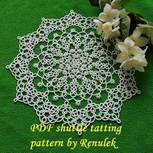 May include: White crochet doily with a circular design, featuring a delicate pattern of loops and knots. The doily is laid on a green surface with white flowers.