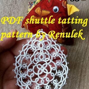 May include: White crocheted doily with a delicate pattern. The doily is held in a hand, with a red and yellow fabric chicken toy in the background. The text "PDF shuttle tatting pattern by Renulek" is visible in the image.