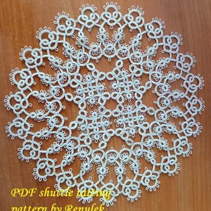 May include: A circular white doily featuring an intricate shuttle tatting lace design with repeating floral and geometric motifs. The delicate needlework creates a complex openwork pattern. Yellow text reads: PDF shuttle tatting pattern by Renulek.