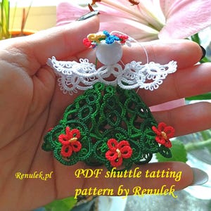 May include: A handmade angel ornament with white lace wings and a green dress adorned with red flowers. The angel has a white head and a colorful halo. The text "PDF shuttle tatting pattern by Renulek" is visible.