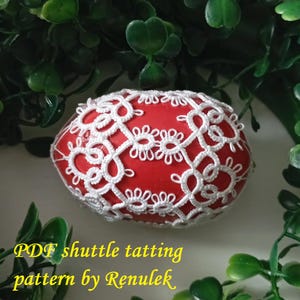 May include: A red egg decorated with white shuttle tatting lace. The egg is surrounded by green foliage. Text on the image reads "PDF shuttle tatting pattern by Renulek."