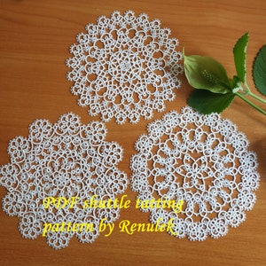 May include: Three white crocheted doilies, each with a delicate floral pattern. The doilies are arranged on a wooden surface. The text "PDF shuttle tatting pattern by Renulek" is visible in the lower part of the image.