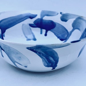 May include: White ceramic bowl with a wide rim, decorated with abstract blue brushstrokes. The bowl's interior and exterior feature a glossy finish, suitable for serving food or as a decorative piece.