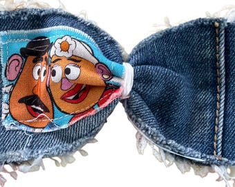May include: A bow-shaped hair accessory crafted from blue denim fabric with frayed edges. The left side features a colorful fabric patch depicting the cartoon characters Mr. and Mrs. Potato Head. The center is cinched with a white elastic band.