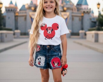 May include: A white short-sleeved t-shirt featuring a red Minnie Mouse silhouette graphic with the name Eve printed below, paired with dark blue denim shorts that have frayed hems and patches of Minnie Mouse imagery, white socks, and white sneakers.