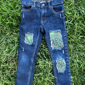 May include: Dark blue denim jeans with distressed patches featuring a green tractor design. The patches are on the knees and thighs of the jeans.