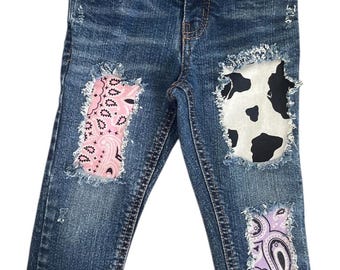 May include: Blue denim jeans with decorative patches. Patches include a pink paisley design, a black and white cow print, and a purple paisley design. The jeans have a classic five-pocket design and a button and zipper closure.