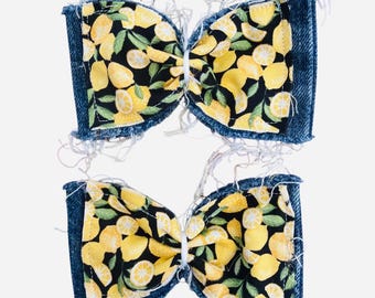 May include: Two bow-shaped hair accessories featuring a black fabric base with a yellow lemon and green leaf pattern. Each bow is bordered by frayed blue denim fabric edges, creating a textured appearance with loose white threads.