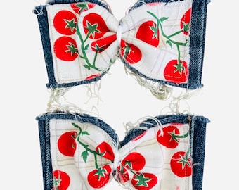 May include: Two hair bows featuring a white fabric with a red tomato and green vine print. The bows are accented with dark blue denim fabric edges that have a frayed, distressed finish with loose white threads along the top and bottom borders.