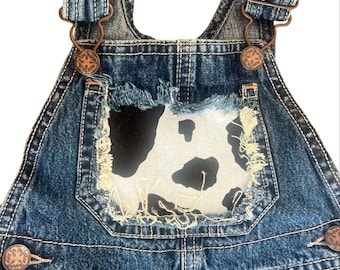 May include: Blue denim overalls with a cow print patch on the chest. The overalls have metal hardware, including adjustable straps and button closures. The denim is distressed with frayed edges around the cow print.