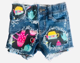 May include: Blue denim shorts with distressed edges. The shorts feature patches with colorful teapot designs. A bow with a teapot print is attached to the waistband. The shorts have a button closure and pockets.