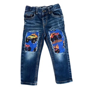 May include: Blue denim jeans with three monster truck patches. The patches feature colorful monster trucks on a blue background. The jeans have a button and zipper closure and are distressed.