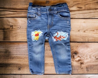 May include: Blue denim jeans with distressed details and patches featuring Winnie the Pooh and Tigger.