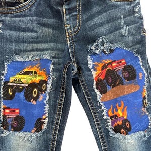 May include: Blue denim jeans with distressed details. Patches of blue fabric feature illustrations of monster trucks engulfed in flames. One truck is green, another red, and a third purple.