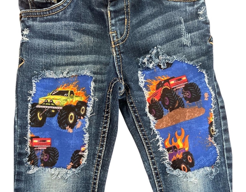 May include: Blue denim jeans with distressed details. Patches of blue fabric feature illustrations of monster trucks engulfed in flames. One truck is green, another red, and a third purple.