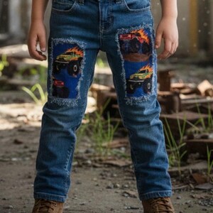 May include: Blue denim jeans with patches featuring monster truck graphics. The patches are on the knees and have a blue background with a distressed edge. The jeans are worn with brown boots.