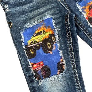 May include: Blue denim jeans with patches featuring monster truck graphics. The patches are blue with a distressed edge, and the monster trucks are in shades of green, red, and orange. The jeans have a classic five-pocket design.