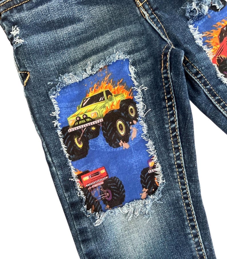 May include: Blue denim jeans with patches featuring monster truck graphics. The patches are blue with a distressed edge, and the monster trucks are in shades of green, red, and orange. The jeans have a classic five-pocket design.