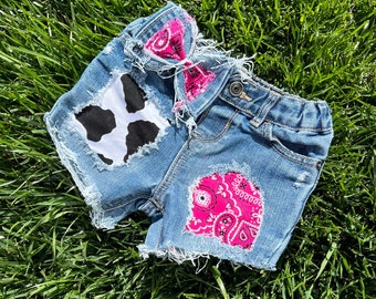 May include: Distressed denim shorts with a pink bandana and a black and white cow print patch. The shorts have frayed edges and are a light blue color.