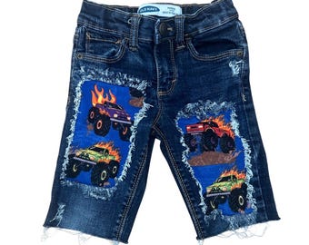 May include: Dark blue denim shorts with distressed edges. The shorts feature patches with monster truck graphics in red, purple, and green, with flames. The shorts are from Old Navy.