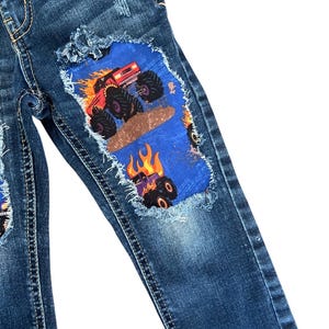 May include: Blue denim jeans with distressed details and patches featuring monster truck graphics. The patches are blue with a monster truck design in red, orange, and black. The jeans have a classic five-pocket design.