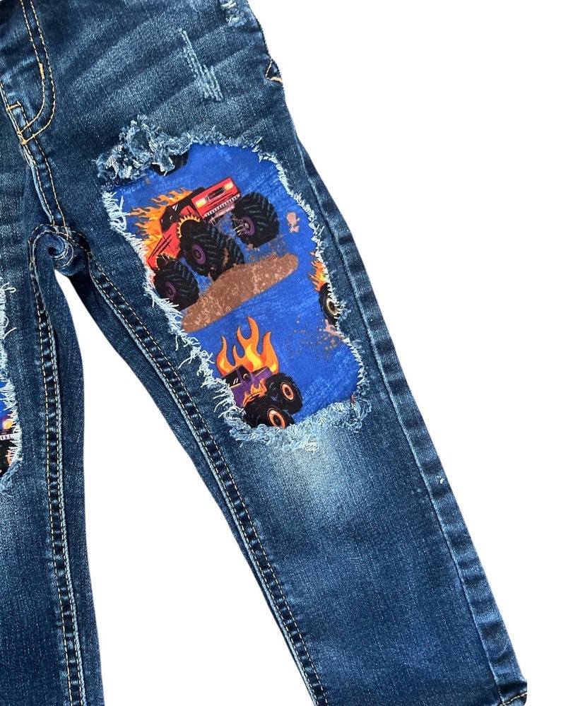 May include: Blue denim jeans with distressed details and patches featuring monster truck graphics. The patches are blue with a monster truck design in red, orange, and black. The jeans have a classic five-pocket design.