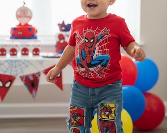May include: A red short-sleeved t-shirt featuring a graphic of Spider-Man with a white web design, paired with blue denim jeans that have rectangular comic book character patches sewn onto the knees and red and blue sneakers.
