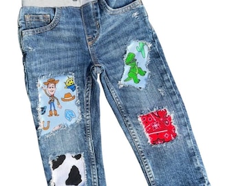 May include: Blue denim jeans featuring a light gray elastic waistband with a drawstring. The pants have distressed patches including a light blue panel with cartoon characters, a green dinosaur, a red bandana print, and a black and white cow print.