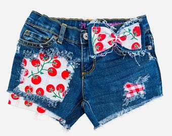 May include: Blue denim shorts featuring frayed edges, a large white patch with a red tomato pattern on the left leg, a smaller red and white checkered patch on the right leg, and a matching white bow with red tomatoes attached to the waistband.