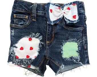 May include: Dark blue denim shorts featuring a frayed hem and distressed areas. One patch is white fabric with a red strawberry print, and another is a green and white gingham check. A matching white strawberry-print fabric bow is attached at the waist.
