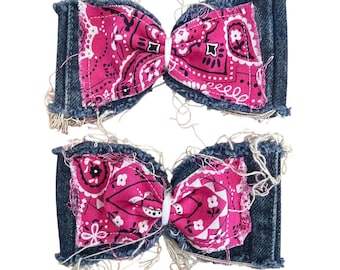 May include: Two hair bows constructed from dark blue denim fabric with frayed edges. Each bow features a central panel of bright pink fabric with a white and black paisley pattern. The bows are cinched in the middle with a white elastic band.