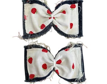May include: Two hair bows featuring a cream-colored fabric with a red strawberry print, layered over dark denim fabric with frayed, distressed edges. The bows are gathered in the center with a thin white band, showcasing a textured, raw-edge finish.