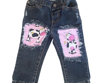 May include: Dark wash denim jeans with a distressed look. Each knee features a pink patch with a cartoon cow illustration. One patch has the word "Moooo" above the cow. The jeans have an elastic waistband.