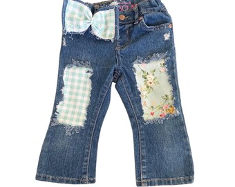 May include: Dark blue denim jeans featuring a frayed light green and white gingham bow at the waist. The legs have distressed patches with a light green and white gingham pattern on the left and a floral print on the right, both with frayed edges.