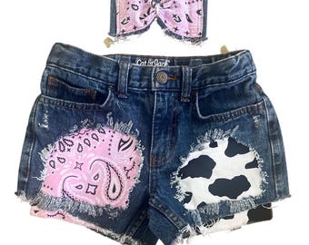 May include: Dark wash denim shorts with distressed edges, featuring pink paisley and cow print patches. A matching pink bow with a paisley pattern is also included. The shorts have the brand name "Cat & Jack" on the waistband.
