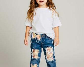May include: A white short-sleeved t-shirt, blue denim jeans with frayed plaid and fox-patterned patches, a matching plaid hair bow, and white sneakers. The jeans feature a distressed look with visible fabric patches on the thighs and shins.