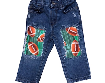 May include: Dark blue denim jeans featuring distressed, frayed knee cutouts that reveal a fabric patch underneath. The patch displays a green football field pattern with white yard lines and multiple brown and white American footballs.