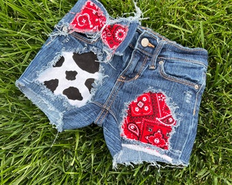 May include: Distressed denim shorts with a cow print patch and red bandana patches. The shorts have frayed edges and a casual look.