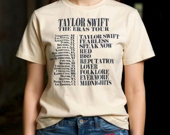 May include: Cream-colored t-shirt featuring the text "TAYLOR SWIFT THE ERAS TOUR" in black. The shirt lists tour locations and album titles. Worn with blue denim shorts with patches and the letters "I TS".