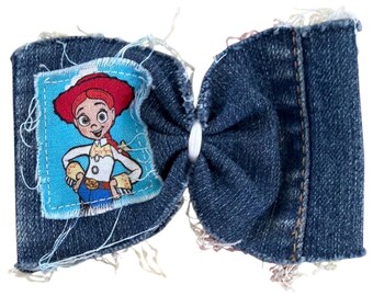 May include: A denim hair bow featuring a frayed edge design and a central white plastic fastener. The left side of the bow displays a rectangular fabric patch with a printed illustration of the character Jessie from Toy Story on a blue background.