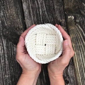 May include: A white rope basket with a woven pattern, held in two hands.