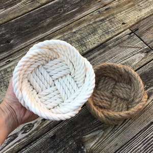 May include: Two round rope baskets, one white and one brown, on a wooden surface. The baskets are made of thick rope and have a woven pattern.