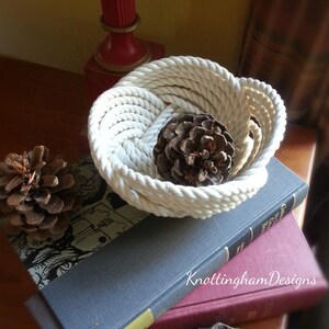 May include: A white rope bowl with a natural pine cone inside. The bowl is sitting on top of two books, one blue and one red. The text "KnottinghamDesigns" is visible on the red book.