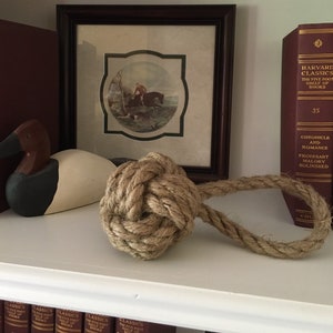 May include: A wooden duck decoy with a brown head and white body sits on a shelf with a brown rope knot and a framed picture of a man riding a horse. The shelf is above a row of books with the text "Harvard Classics The Five Foot Shelf of Books" on the spine.