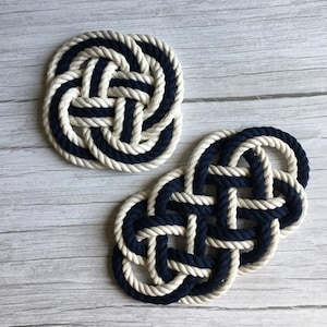 May include: Two rope coasters, one round and one shaped like a figure of eight, are made with white and navy blue rope. The coasters are on a light wood surface.