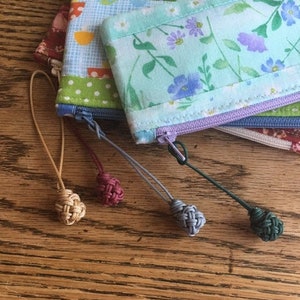 May include: Four small zippered pouches with fabric patterns and colorful tassels. The pouches are made of different fabrics with floral, polka dot, and geometric patterns. The tassels are made of different colors of cord.