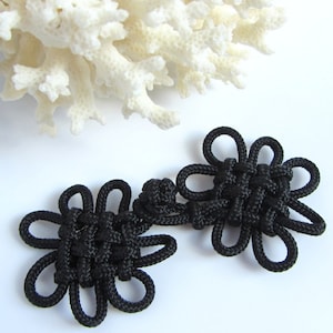 May include: Two black Chinese knot buttons in the shape of a flower. The knots are tied with a thick black cord and have a woven pattern.