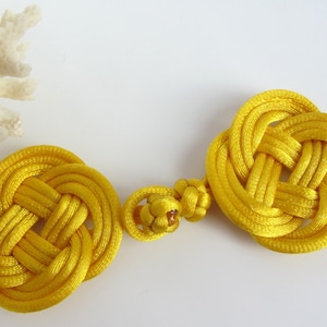 May include: Two bright yellow, intricately knotted decorative elements, possibly cord or rope, are displayed against a white background. The knots are in a circular pattern, and a small, connecting knot joins them. A piece of white coral is in the background.