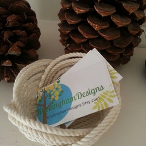 May include: A white rope bowl containing a business card that reads "Knottingham Designs" and "KnottinghamDesigns.Etsy.com". The bowl is sitting on a white surface with two brown pine cones.
