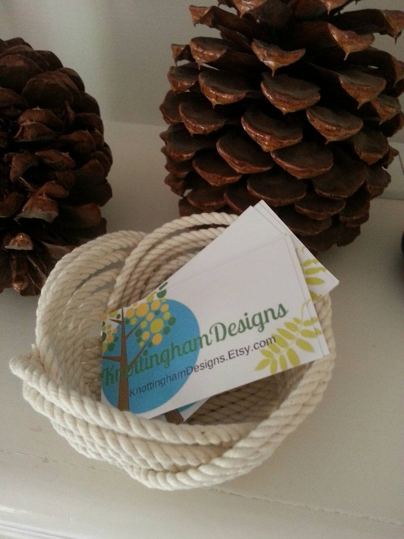 May include: A white rope bowl containing a business card that reads "Knottingham Designs" and "KnottinghamDesigns.Etsy.com". The bowl is sitting on a white surface with two brown pine cones.